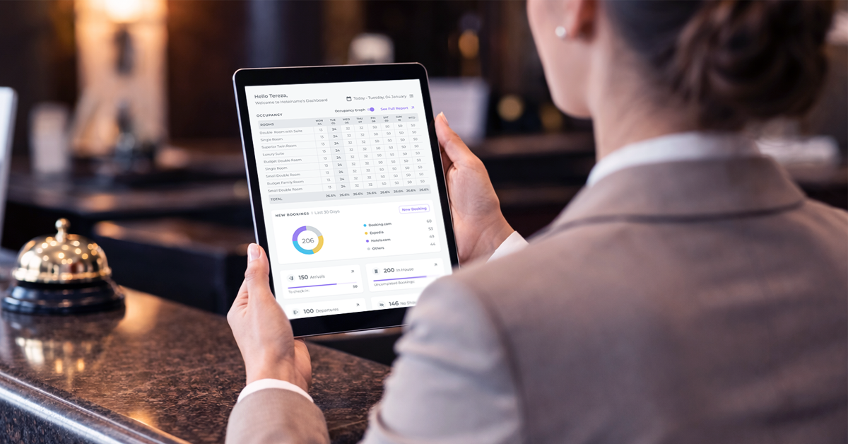 A hotel receptionist holding a tablet with a PMS interface open.